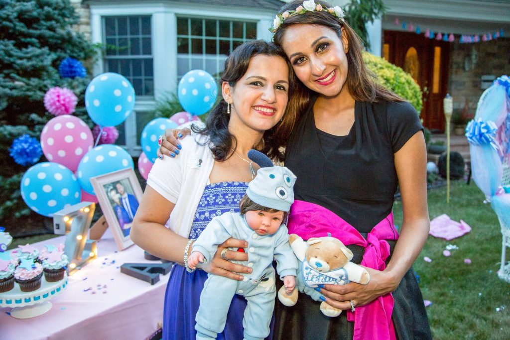 expecting mother withe garland and teddy bear smiling with her fried who is holding a baby doll for the gender reveal party