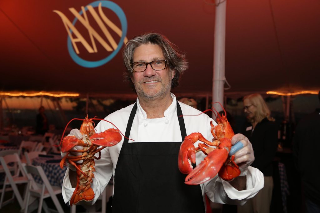 man with medium long hair and glasses smiling as he holds up two lobsters